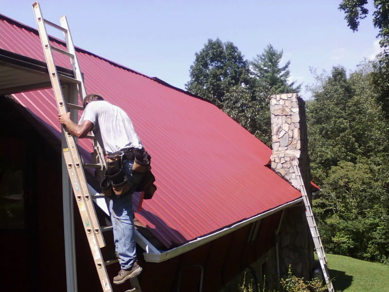 Roofer on a ladder beside a red metal roof and stone chimney — Soffit & Fascia Repair by Roofing Brothers in Whigham